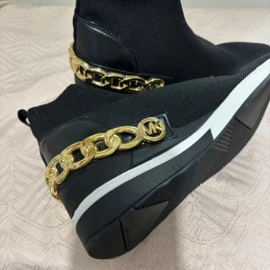 Svarta Michael Kors boots med guldkedja - Svarta boots från Michael Kors i stickad textil med detaljer i skinn. Snygg guldfärgad kedja och MK-logga bak på hälen. Vit rand på sulan ger en sportig touch. Perfekta för dig som vill sticka ut med stilrena detaljer.