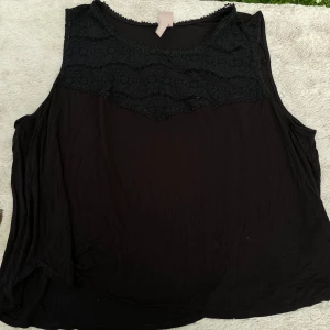 Svart ärmlös topp med spets från H&M - Svart ärmlös topp från H&M i storlek 3XL. Toppen har en snygg spetsdetalj över bröstet och en mjuk, flowy passform. Perfekt för dig som gillar en enkel men ändå cool look. Materialet känns lätt och bekvämt mot huden.