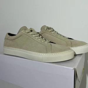 Beige mockasneakers från Les Deux - Stilrena sneakers i beige mocka från Les Deux. Skorna har klassisk låg profil, snörning och vit platt sula. Perfekta för dig som gillar minimalistisk och clean stil. Passar till det mesta och ger en fräsch vibe till outfiten. Helt nya, perfekta för sommaren.