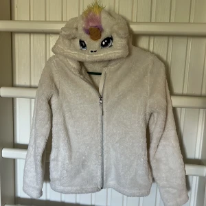 Vit teddyjacka enhörning H&M 134/140 - Supermjuk vit teddyjacka från H&M med dragkedja och huva som har ett gulligt enhörningsansikte, öron och färgglad man i lila, gult och grönt. Jackan är fluffig och har långa ärmar.