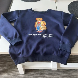 Blå Polo Bear tröja från Ralph Lauren - Säljer en mörkblå crewneck från Polo Ralph Lauren med ikoniska Polo Bear-trycket framtill. Tröjan har rund hals, ribbade muddar och är i mjuk bomullsmix. Perfekt för dig som gillar streetstyle och vill sticka ut med en klassisk men lekfull look. Vid några funderingar hör av dig// William 