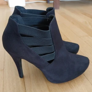 Svarta stövletter med klack och resår - Snygga svarta stövletter i mocka-look med höga klackar och breda elastiska band på sidorna. Skorna har spetsig tå och en stilren design som passar perfekt till både jeans och klänning.