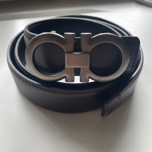 Brunt skärp från Salvatore  - Snyggt brunt bälte med ikoniskt dubbel-Gancini-spänne i matt silver. Bältet är gjort i äkta skinn med diskreta sömmar längs kanterna. Perfekt accessoar för att lyfta din outfit med en lyxig touch.