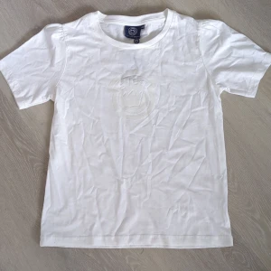 Vit t-shirt med pufftryck från Svea - Oanvänd vit t-shirt från Svea med stilrent pufftryck av loggan på bröstet. Klassisk rund halsringning och korta ärmar. Tillverkad i 100% bomull för en skön och luftig känsla. Perfekt för dig som gillar enkel och clean stil.