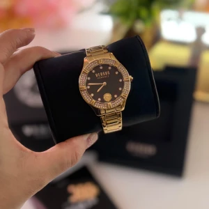 Guldig Versus Versace klocka med stenar - Snygg klocka från Versus Versace i guldigt metallarmband och svart urtavla med diskret randigt mönster. Urtavlan är dekorerad med gnistrande stenar runt ramen och lejonlogga vid sexan. Perfekt accessoar för att sticka ut och ge looken extra lyx.