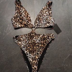Leopardmönstrad baddräkt från South Beach, mer som strl 40 - Snygg leopardmönstrad baddräkt från South Beach med cut-out detaljer och ring framtill. Justerbara axelband och coolt högt skuren modell. Perfekt för dig som vill sticka ut på stranden! Liten i storlek och oanvänd.