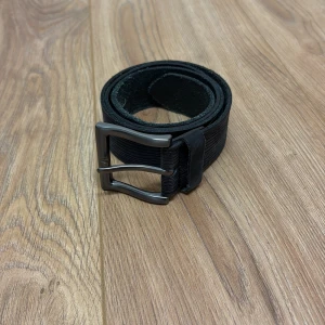 Svart randigt skärp i skinn - Snyggt svart bälte i skinn med diskret randig struktur längs hela remmen. Bältet har ett stilrent metallspänne i mörkgrått som ger en modern touch. Perfekt accessoar för att lyfta din outfit med en enkel men cool detalj.