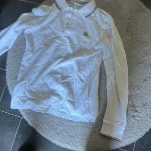 Vit långärmad pikétröja Moncler - Vit långärmad pikétröja från Moncler med klassisk krage och knäppning framtill. Tricolore-detaljer på krage och ärmslut samt broderad Moncler-logga på bröstet. Tillverkad i bomull för en skön och stilren look.