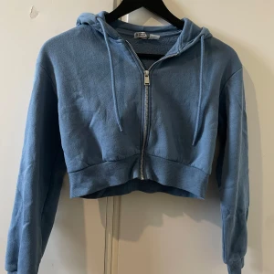 Blå croppad hoodie med dragkedja - Säljer en blå croppad hoodie från NICO med dragkedja framtill och snörning i huvan. Tröjan har långa ärmar, ribbade muddar och är gjord i mjuk bomullsmix. Perfekt för en chill och avslappnad stil.