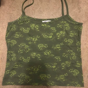Grönt linne - Snygg grön linnetopp med tunna axelband. Toppen har ett blommigt mönster i ljusare grönt som ger en fräsch och somrig vibe. Perfekt att matcha med jeans eller kjol för en chill look. Materialet känns mjukt och stretchigt.