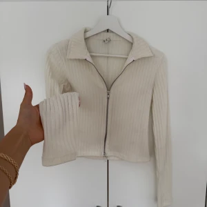 Ribbad beige kofta med dragkedja - Säljer en ribbad beige kofta med lång ärm och krage. Tröjan har en hel dragkedja framtill och är i ett stickat material som känns både mjukt och stilrent. Perfekt till jeans eller kjol för en clean look.