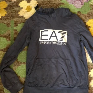 Mörkblå EA7 hoodie från Emporio Armani - Snygg mörkblå hoodie från Emporio Armani EA7 med stor logga framtill. Tröjan har huva, lång ärm och en klassisk känguruficka. Perfekt för dig som gillar streetwear och vill ha en stilren look med premiumkänsla.