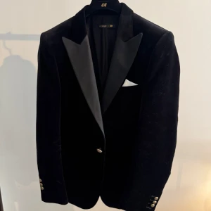 Svart sammetskavaj Balmain x H&M - Exklusiv svart sammetskavaj från Balmain x H&M med breda slag i satin och snygga guldknappar vid ärmslut. Klassisk passform och elegant design, perfekt för dig som vill sticka ut med stil. Ficka med vit näsduk för extra detalj. 