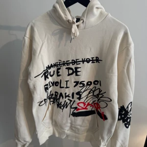 Vit hoodie med graffiti från Manière De Voir - Säljer en vit hoodie från Manière De Voir med coolt graffitiinspirerat tryck i svart och rött både fram och bak. Tröjan har huva med snörning och känguruficka. Perfekt för dig som gillar streetstyle och unika plagg.