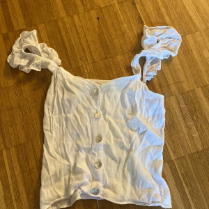 Vit linnetopp med volangaxelband - Supersöt vit linnetopp med volangiga axelband och knappar framtill. Toppen har en smockad rygg för extra komfort och sitter snyggt på kroppen. Perfekt för varma dagar och ger en fräsch och somrig look.