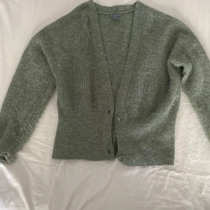 Grön stickad kofta från Lindex - Mysig ljusgrön stickad kofta från Lindex med v-ringning och två knappar framtill. Koftan har långärmad passform och är ribbstickad för en chill och trendig look. Perfekt att slänga över en t-shirt eller linne när det är lite kyligt ute.