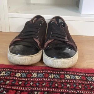 Vinröda sneakers från Lanvin med lackad tå - Snygga bruna sneakers från Lanvin med svart lackad tå och vita sulor. Ovandelen är i mocka med svarta skosnören och bruna detaljer runt öppningen. Klassisk rund tå och platt sula ger en clean look som funkar till det mesta.