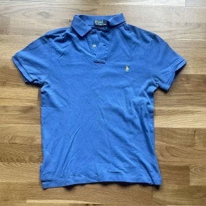 Ralph Lauren piké  - Klassisk blå pikétröja från Ralph Lauren med gul broderad logga på bröstet. Tröjan har korta ärmar, krage och knäppning med två knappar. Tillverkad i bomull för en skön och luftig känsla. Perfekt för en avslappnad men ändå stilren look.