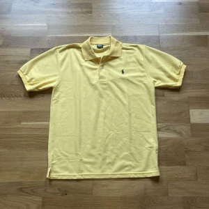 Ralph Lauren piké  - Gul pikétröja från Polo Ralph Lauren med klassisk krage, två knappar och broderad logga på bröstet. Tillverkad i mjuk bomull och har korta ärmar. Perfekt för dig som gillar stilren och sportig look. I fint skick men lite blekt färg 