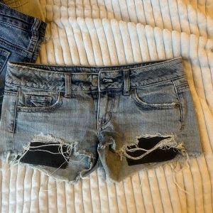 Lågmidjade jeansshorts från American Eagle - Snygga ljusblå jeansshorts från American Eagle med låg midja och råa, fransiga kanter. Shortsen har slitna detaljer och trasiga partier framtill för en cool, avslappnad vibe. Klassisk femficksmodell med knappar och dragkedja. Perfekta för sommaren! Storlek 00 eller Xs