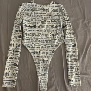 Vit långärmad body med svart text - Trendig vit body med lång ärm och svart textprint i olika typsnitt över hela plagget. Hög skärning vid höften och rund halsringning. Perfekt för dig som vill sticka ut med en edgy och cool stil.