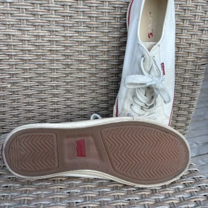 Vita Levi's sneakers med röd detalj - Klassiska vita sneakers från Levi's med röd logga på sidan och röd rand längs sulan. Skorna har snörning och en platt, brun sula i gummi. Perfekta för en clean och avslappnad stil.
