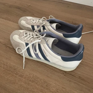 Adidas Gazelle vita och blå sneakers - Snygga Adidas Gazelle sneakers i vitt och blått med klassiska tre ränder på sidan. Ovandel i mocka och skinn, med vit sula och snörning. Perfekta för dig som gillar retrostil och vill ha ett par ikoniska sneakers i garderoben.