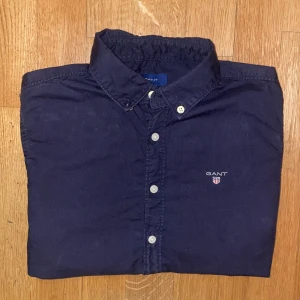 Mörkblå skjorta från gant. - Klassisk mörkblå skjorta från GANT med vit knappstängning och broderad logga på bröstet. Skjortan har lång ärm och stilren krage, perfekt för en clean och snygg look. Tillverkad i mjuk bomull som känns skön mot huden.