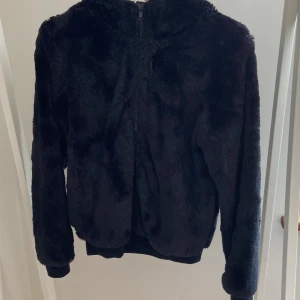 Svart teddyjacka med huva från KappAhl - Mysig svart teddyjacka från KappAhl med huva och dragkedja framtill. Jackan har långärmade ärmar och är tillverkad i mjukt, fluffigt material som håller dig varm. Perfekt för kyliga dagar när du vill ha en chill och snygg look.