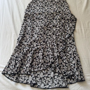 Blommig volangkjol från lager 157 - Supersöt svart kjol med vita och beige blommor från lager 157. Kjolen har volangdetaljer och omlottkänsla, perfekt för dig som gillar en feminin och flowy stil. Resår i midjan för extra komfort.