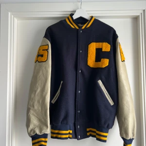 Vintage bomberjacka Coronado Band - Säljer en klassisk vintage Amerikansk fotboll varsity bomberjacka från Coronado Band, New Mexico State university NMSU i USA, tillverkad av Maple. Jackan har mörkblå ullig kropp, kontrasterande beige skinnärmar och gula detaljer. Broderade patches på ärmar och rygg, samt randiga muddar i gult och blått. Perfekt för dig som gillar retro college-stil. OBS! Det finns ett hål i vänster armhåla, det påverkar inte slitaget och syns inte från utsidan. Jag kan skicka en bild om det behövs.