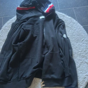 Svart Moncler hoodie med dragkedja - Svart hoodie från Moncler med hel dragkedja, fickor fram och huva med röd, vit och blå detalj. Klassisk Moncler-logga på ärmen. Tillverkad i mjuk bomull, perfekt för chill dagar eller när du vill ha en clean streetstyle-look.