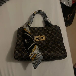 Mönstrad handväska med scarf-detalj - Snygg handväska i mörkbrunt med små guldiga mönster och guldfärgat lås framtill. Handtaget är inslaget i en silvrig scarf med svarta och gula detaljer som ger väskan en unik touch. Perfekt storlek för det viktigaste och riktigt trendig look.