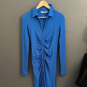 Blå ribbad klänning från NA-KD - Snygg blå ribbad klänning från NA-KD med lång ärm, krage och knappar hela vägen framtill. Klänningen har en cool rynkad detalj i mitten som ger en unik look. Perfekt för dig som gillar trendiga och bekväma plagg med lite edge.