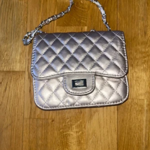Quiltad silver axelväska med kedja - Säljer en snygg quiltad axelväska i metallic silver med kedjerem. Väskan har ett klassiskt rutmönster, vridlås framtill och ett fack med dragkedja inuti. Perfekt storlek för det viktigaste och ger en lyxig touch till din outfit.
