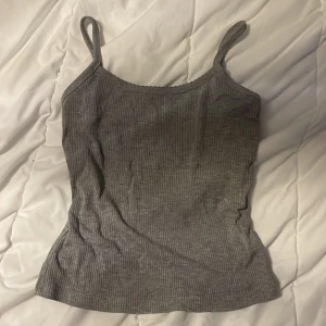 Grå ribbad linnetopp - Säljer en enkel och stilren grå ribbad linnetopp med smala axelband. Toppen har en lätt vågig kant upptill och är i mjukt, stretchigt material som sitter skönt på kroppen. Perfekt att matcha med jeans eller kjol för en avslappnad look.