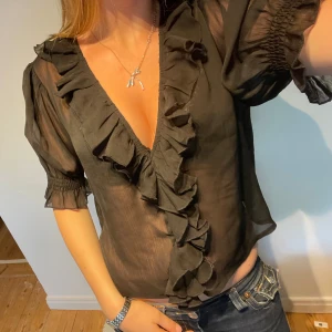Svart volangblus från Boohoo - Säljer en svart, transparent blus från Boohoo med djup v-ringning och stora volangdetaljer längs halsringningen. Blusen har korta puffärmar med resår och är tillverkad i ett lätt, luftig material som ger en snygg och trendig look. Står eu 42 men passar mig som vanligtvis är en S i storlek