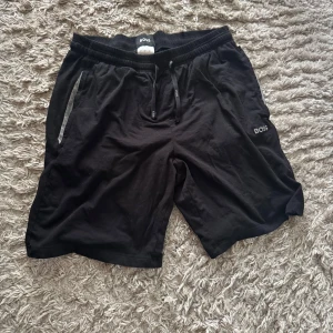 Svarta shorts från BOSS med snörning - Svarta shorts från BOSS med broderad logga på benet. De har elastisk midja med snörning och praktiska fickor med en diskret grå detalj. Perfekta för chill dagar eller träning, tillverkade i mjuk bomull för maximal komfort.