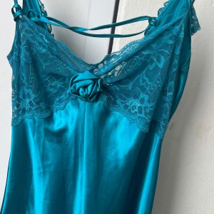 Turkos långklänning med spets och rosett - Snygg turkos lång natlinen i satinliknande material med spetsdetaljer upptill och längs ryggen. Klänningen har smala axelband, dekorativ rosett framtill och volang med spets bak. Onesize