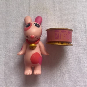🐈 - Bilderna visar en söt figur med kaninöron, stora ögon och rosa fläckar, samt en burk märkt 'Cat Food'. Figuren bär en klocka runt halsen och har små vingar på ryggen. Ingen bok syns på bilderna.