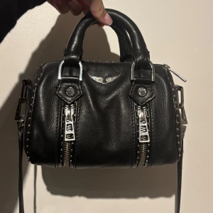 Zadig voltaire Sunny Nano Studs bag -  svart handväska från Zadig & Voltaire i skinn med silvriga metalldetaljer, dubbla handtag och avtagbar axelrem. Väskan har dekorativa dragkedjor framtill, nitar längs kanterna och ett litet emblem med vingar. Minsta modellen