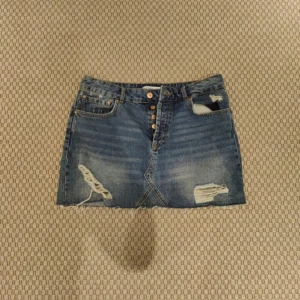 Blå jeanskjol med slitningar från Zara  - Snygg blå jeanskjol med slitna detaljer och rå kant nertill. Kjolen har knappar framtill och klassiska fickor både fram och bak. Perfekt för dig som gillar en avslappnad och trendig stil.