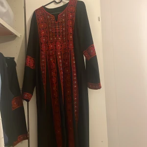 Svart långklänning med röd brodyr - Unik svart långklänning med fantastiska röda broderier och detaljer i geometriska mönster. Klänningen har långa ärmar och v-ringad hals. Broderierna har inslag av små färgdetaljer i blått och vitt. Perfekt för dig som vill sticka ut med en traditionell och färgstark look. Har en annan likadan i storlek S
