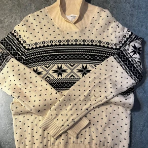 Beige och svart mönstrad tröja Selected Homme - Snygg beige stickad tröja från Selected Homme med svart nordiskt mönster och stjärndetaljer. Tröjan har lång ärm, ribbad krage och muddar. Perfekt för kalla dagar när du vill ha en stilren och mysig look.