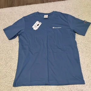 Blå Champion t-shirt i bomull - Snygg blå t-shirt från Champion med klassisk logga broderad på bröstet och ärmen. T-shirten har rund halsringning och är tillverkad i mjuk bomull. Perfekt basic-plagg med stilren look och normal passform.