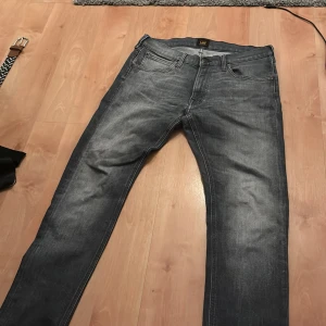 Grå jeans från Lee - Snygga grå jeans från Lee med klassisk femficksdesign och diskret tvättad look. Jeansen har normal passform och raka ben, perfekt för en stilren vardagsoutfit. Tillverkade i mjukt jeanstyg med Lee-logga på knappen och bak i linningen.