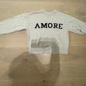 Beige stickad tröja med AMORE-print - Superfin beige stickad tröja från Gina med svart text 'AMORE' framtill. Tröjan har rund hals, långa ärmar och breda ribbade muddar vid ärmslut och nederkant. Perfekt för dig som gillar enkel men snygg stil. PRIS KAN DISKUTERAS🩷🩷