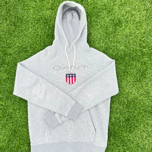 Grå hoodie från GANT med logga - Snygg grå hoodie från GANT med broderad logga och sköld på bröstet. Tröjan har huva med vita snören och en klassisk känguruficka framtill. Perfekt för en chill och sportig look. Tillverkad i mjuk bomullsmix för extra komfort.