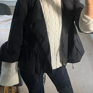 Svart overshirt med volangdetalj - Snygg svart overshirt med bred krage och volangdetalj nedtill. Jackan har knytband i midjan och en loose passform som ger en chill vibe. Perfekt att slänga över en stickad tröja för en cool lager-på-lager-look. Tunn och perfekt till sommaren 