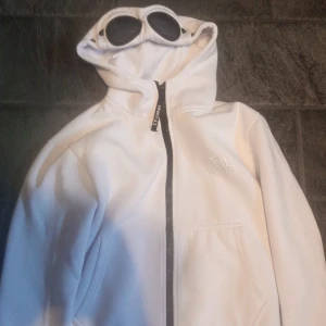 Vit C.P. Company goggle zip M - Säljer en vit goggle hoodie från C.P. Company i storlek M. Hoodien har huva med ikoniska goggles, dragkedja framtill och broderad logga på bröstet. Snygg och clean design.